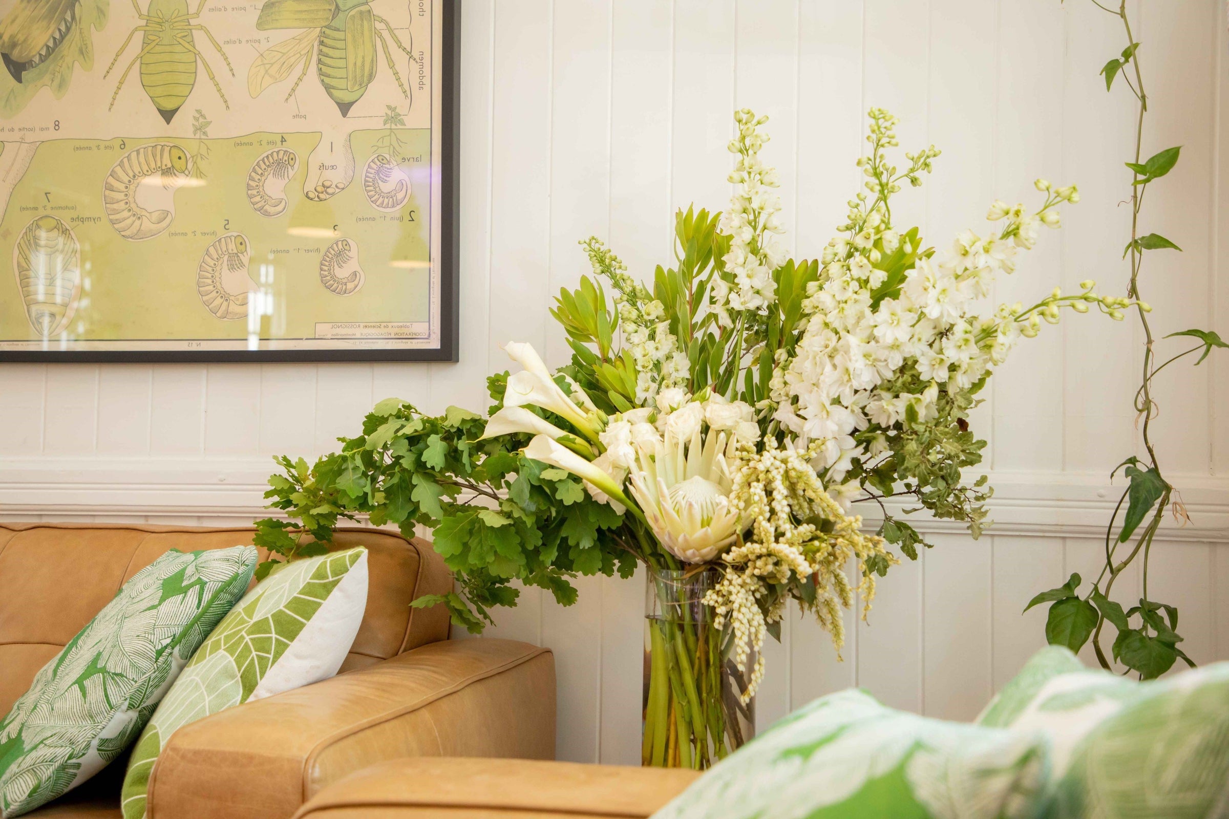Large glass vase arrangement filled with white and green flowers and foliage placed in room with tan coloured lather lounges