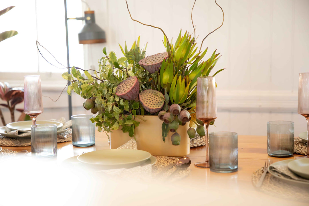Side view of lemon coloured ceramic hedge vase filled with Earthy foliage lotus pods and gum nuts  surrounded by a muted table setting