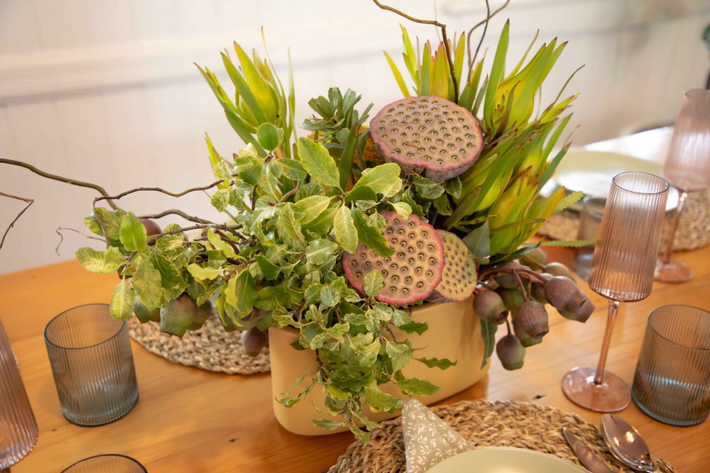 Closer view of lemon coloured ceramic hedge vase filled with Earthy foliage lotus pods and gum nuts  surrounded by a muted table setting