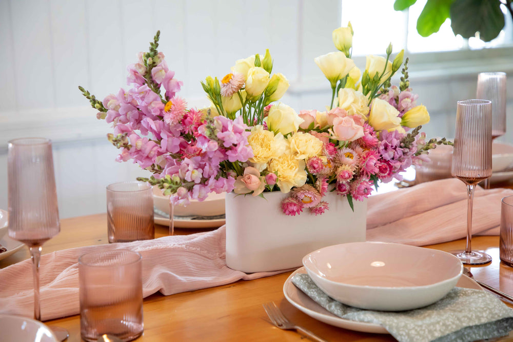 Off white ceramic hedge vase spilling with pastel coloured flowers in pink, lemon and mauve set on pink table settings