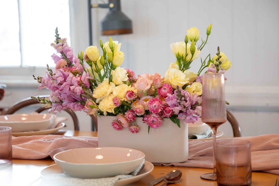 Side view of off white ceramic hedge vase spilling with pastel coloured flowers in pink, lemon and mauve set on pink table settings