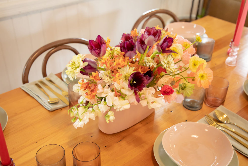 Over view of ceramic hedge vase spilling with Meadow style flowers in peach, purple, tangerine, light pink and sunshine yellow on muted table setting