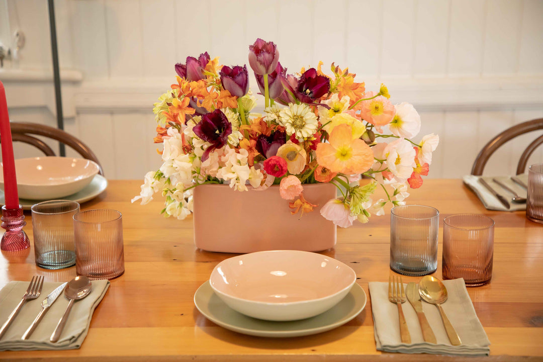 Side view of ceramic hedge vase spilling with Meadow style flowers in peach, purple, tangerine, light pink and sunshine yellow on muted table setting