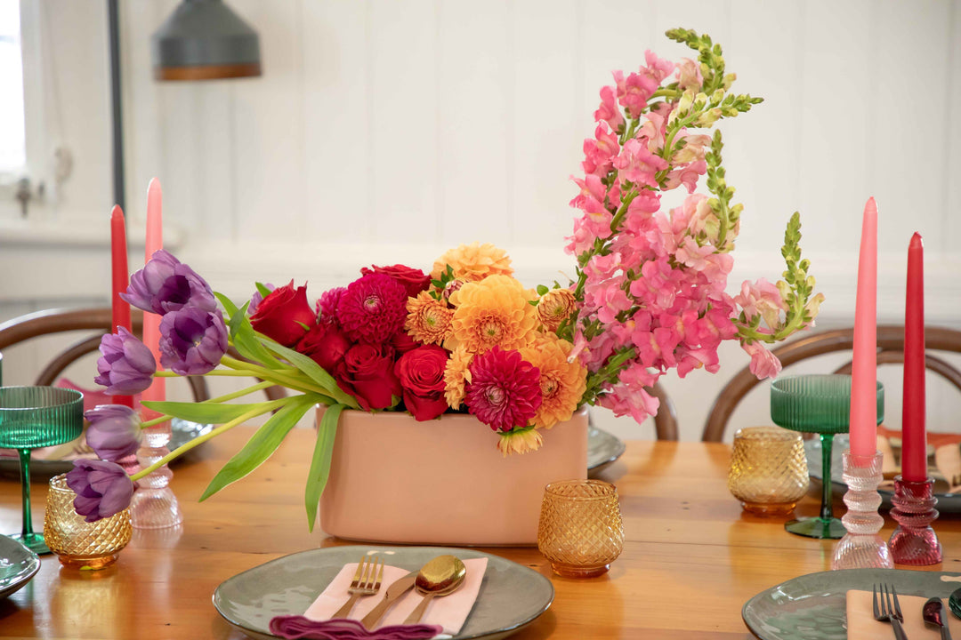 Front view of light pink ceramic hedge vase filled with bright bold overflowing flowers surrounded by a bright table setting