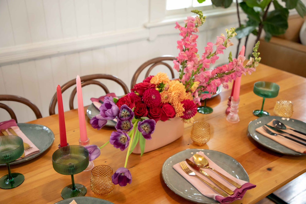 Overview of light pink ceramic hedge vase filled with bright bold overflowing flowers surrounded by a bright table setting
