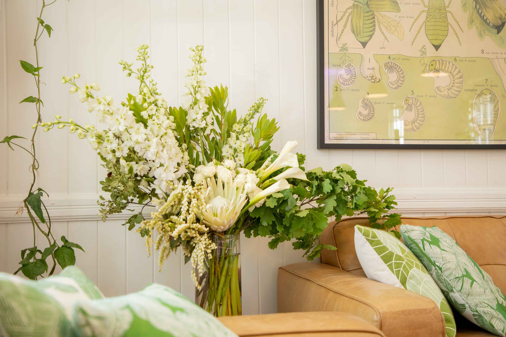 Large vase arrangement filled with green and white flowers and foliage white king protea white delphinium placed in glass vase 