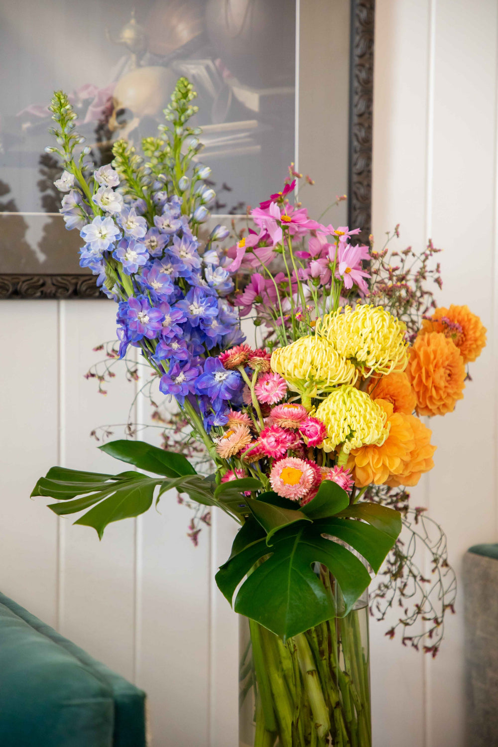 Large vase arrangement filled with bright bold flowers blue delphinium yellow waratah orange dahlia pink cosmo and strawflowers in glass vase 