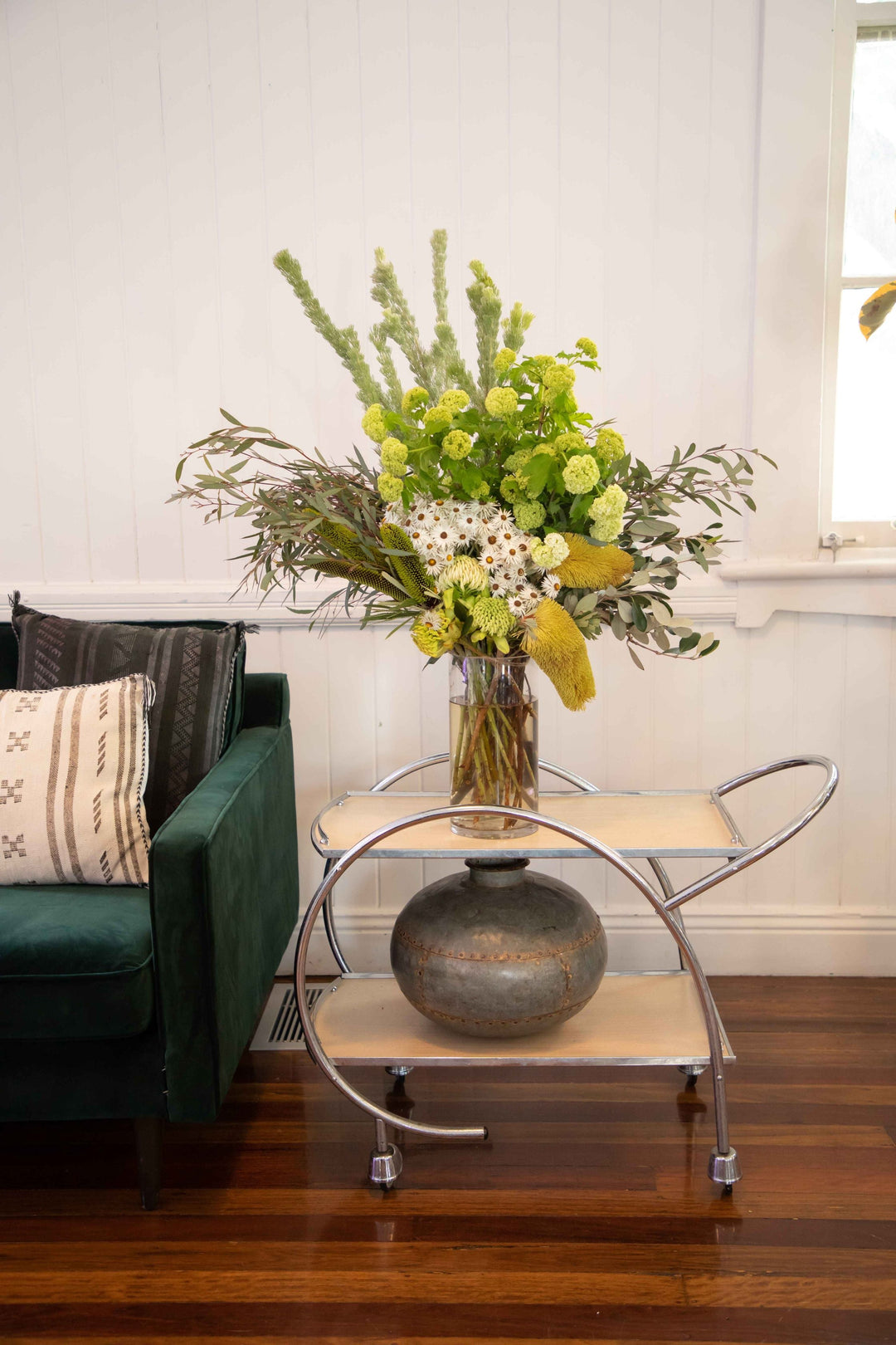 Large vase arrangement filled with green foliage and flowers placed in glass vase sitting on top of vintage silver  drinks card