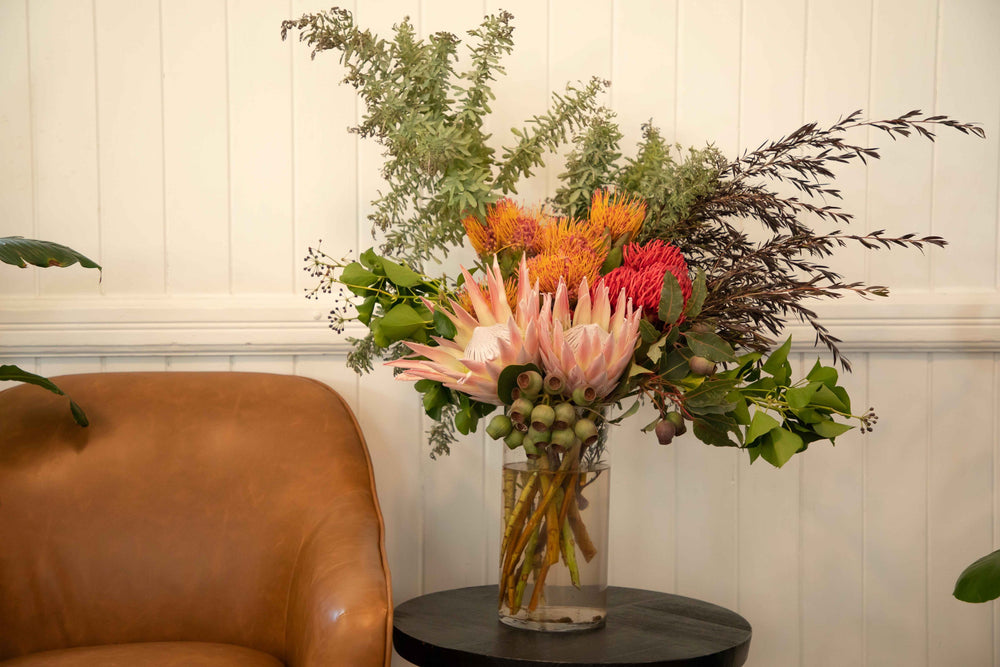 Large vase arrangement filled with Australian and South African native flowers and foliage pink king protea pincushions gum nuts placed in glass vase sitting on wooden side table