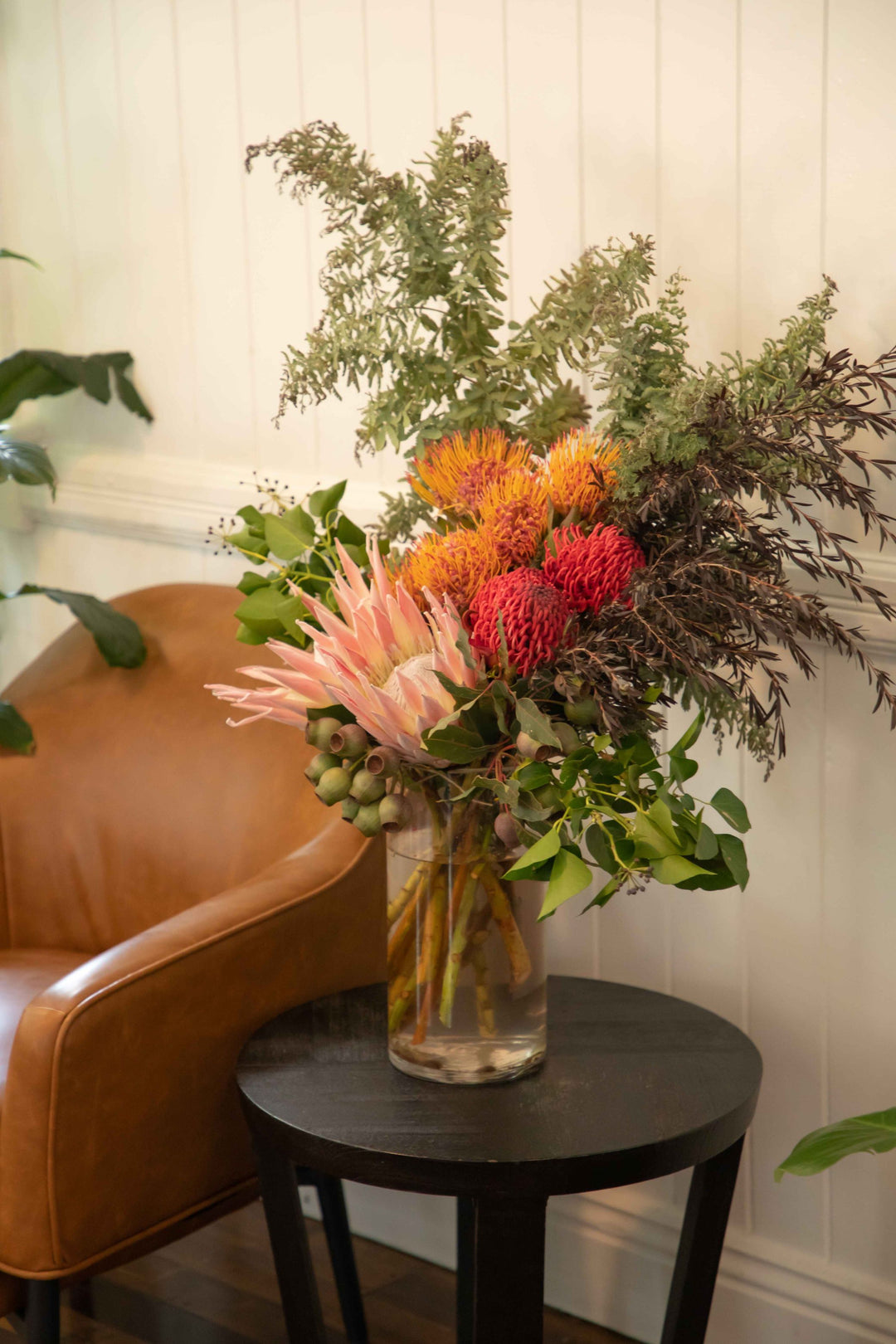 Large vase arrangement filled with Australian and South African native flowers and foliage pink king protea pincushions gum nuts placed in glass vase sitting on wooden side table